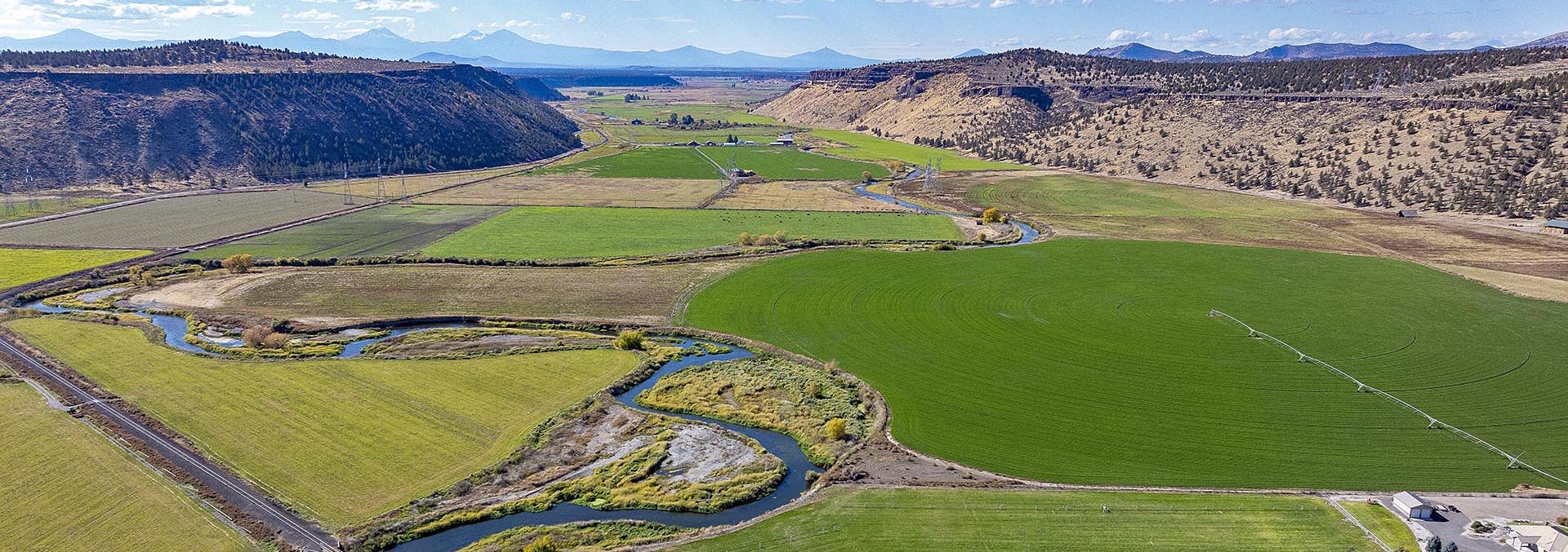 Aerial view of green fields and a winding river through a valley framed by rugged hills and distant mountains under a blue sky.",