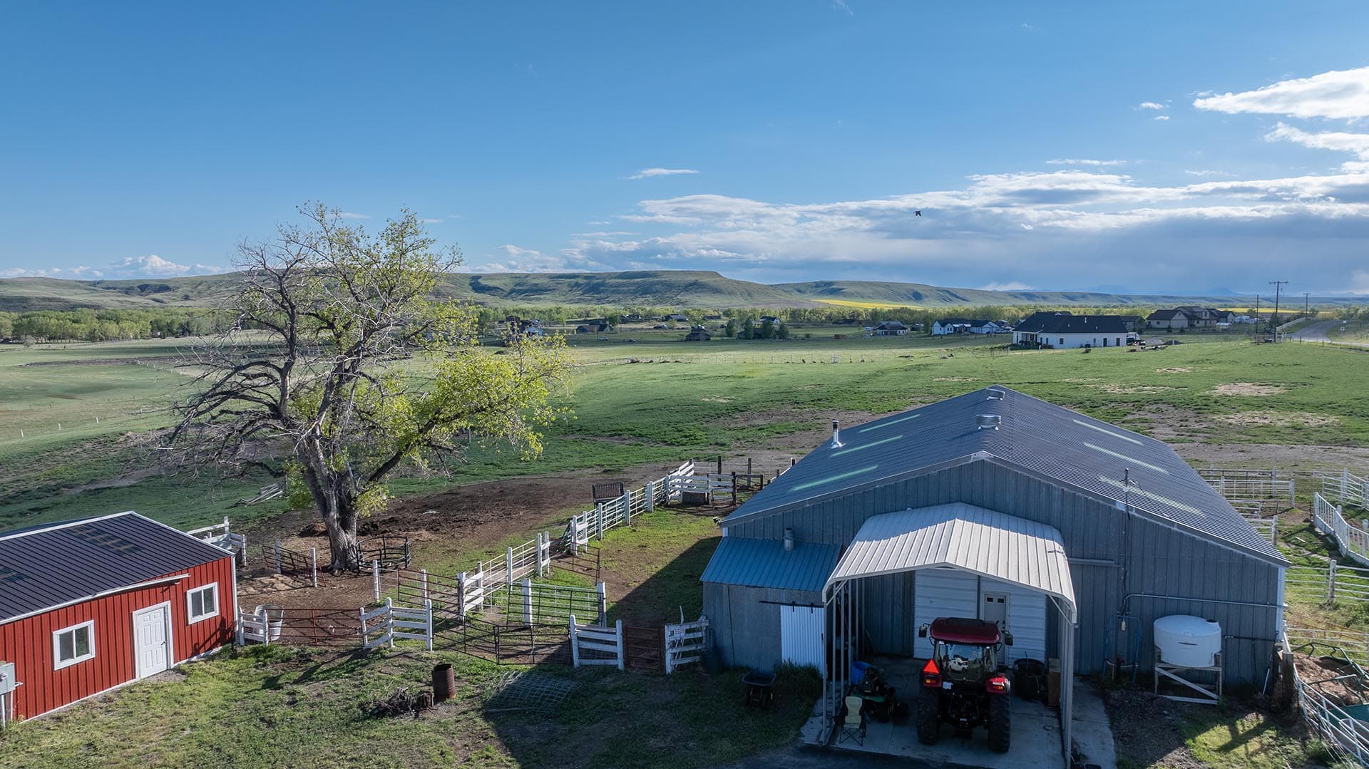 Wide rural farm scene with red barn, metal-roofed outbuilding, fenced pastures, and distant hills under a blue sky.