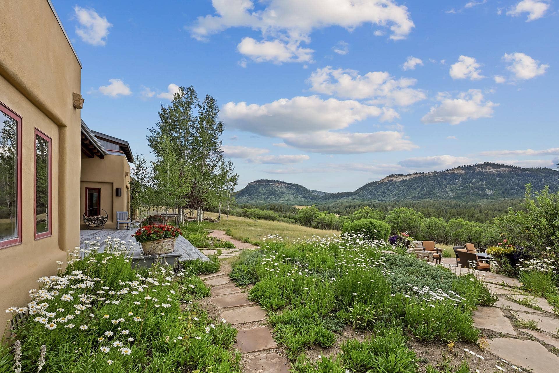 Adobe-style house with red-trim windows and a flower-filled courtyard leading to rolling hills and distant mountains.