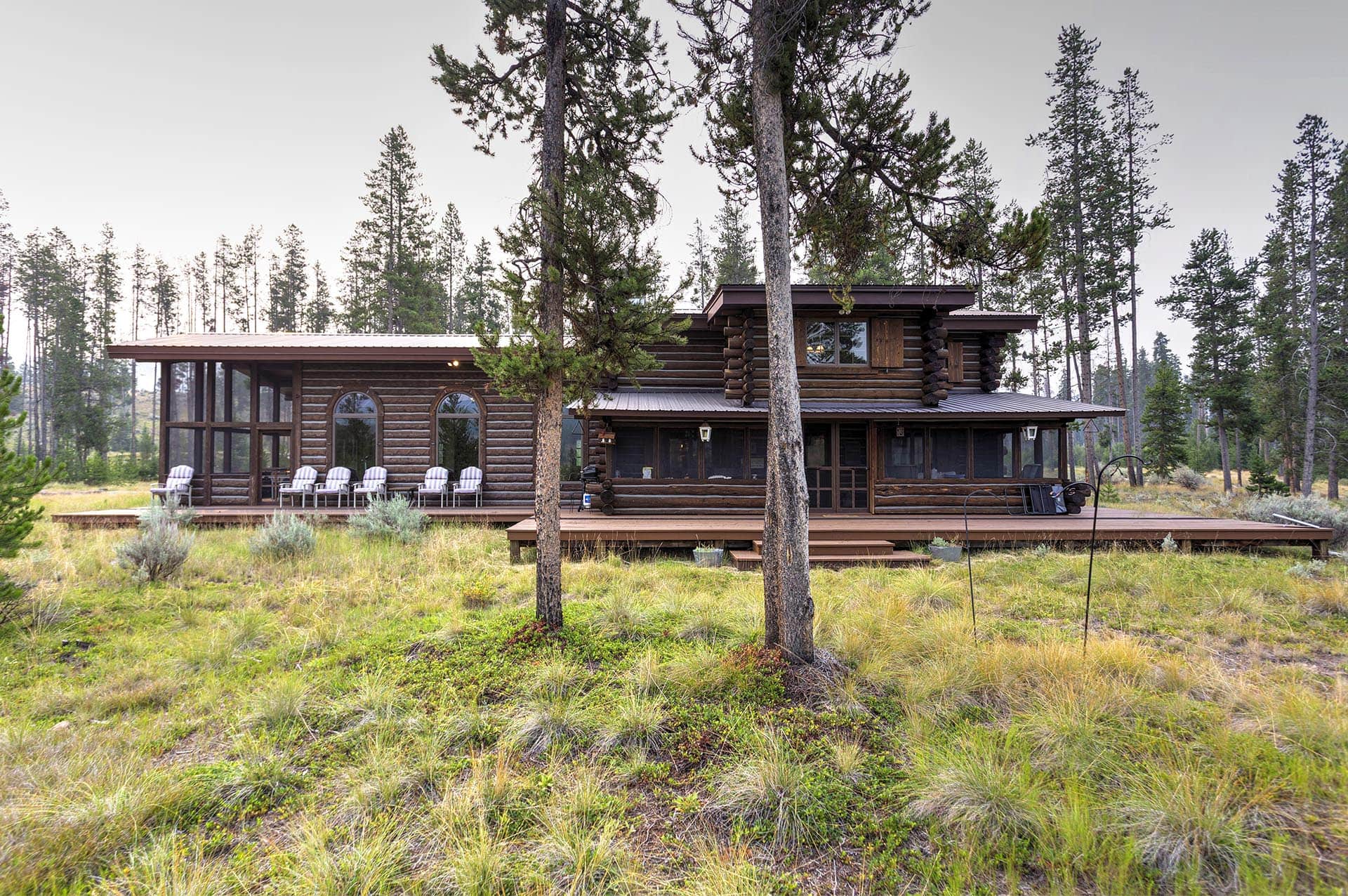 Log hillside log cabin with a wooden deck and several white outdoor chairs, surrounded by trees in a forest clearing.