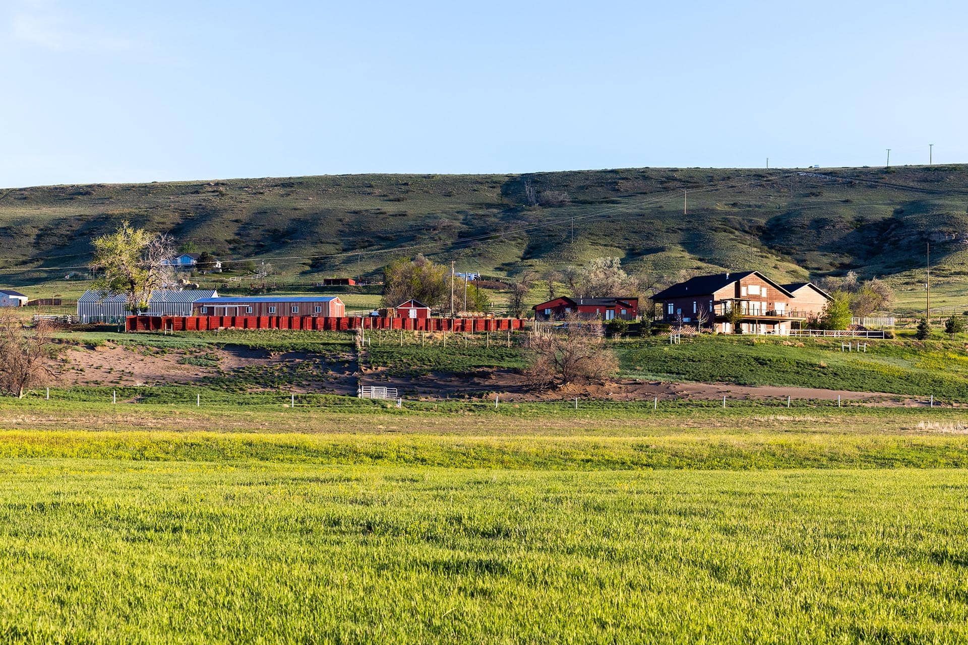 Rural farmstead with red outbuildings and a long red fence at the base of a grassy hill under a clear blue sky.