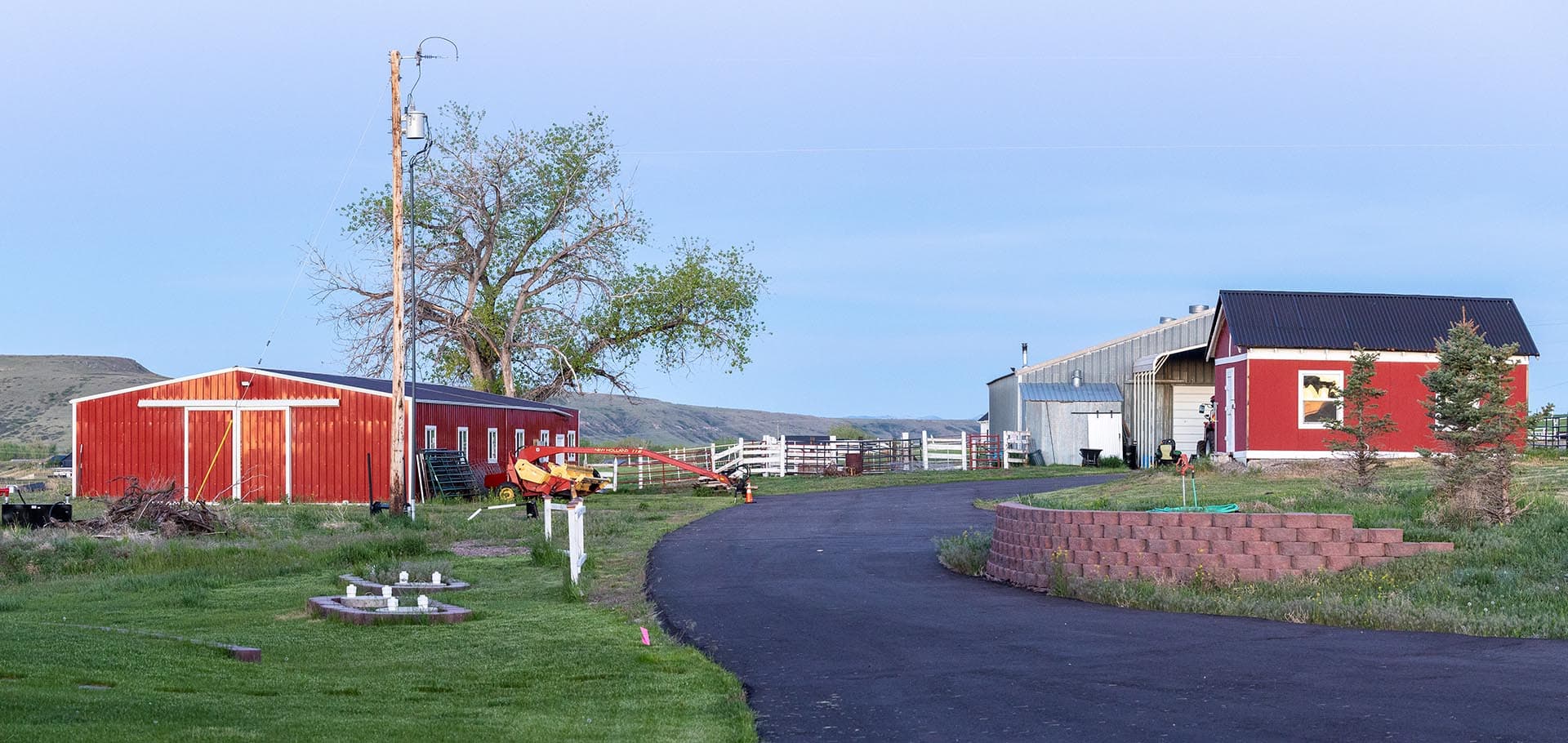 Red barn and red outbuilding on a rural farm with a curved asphalt driveway and green grass under a clear blue sky.