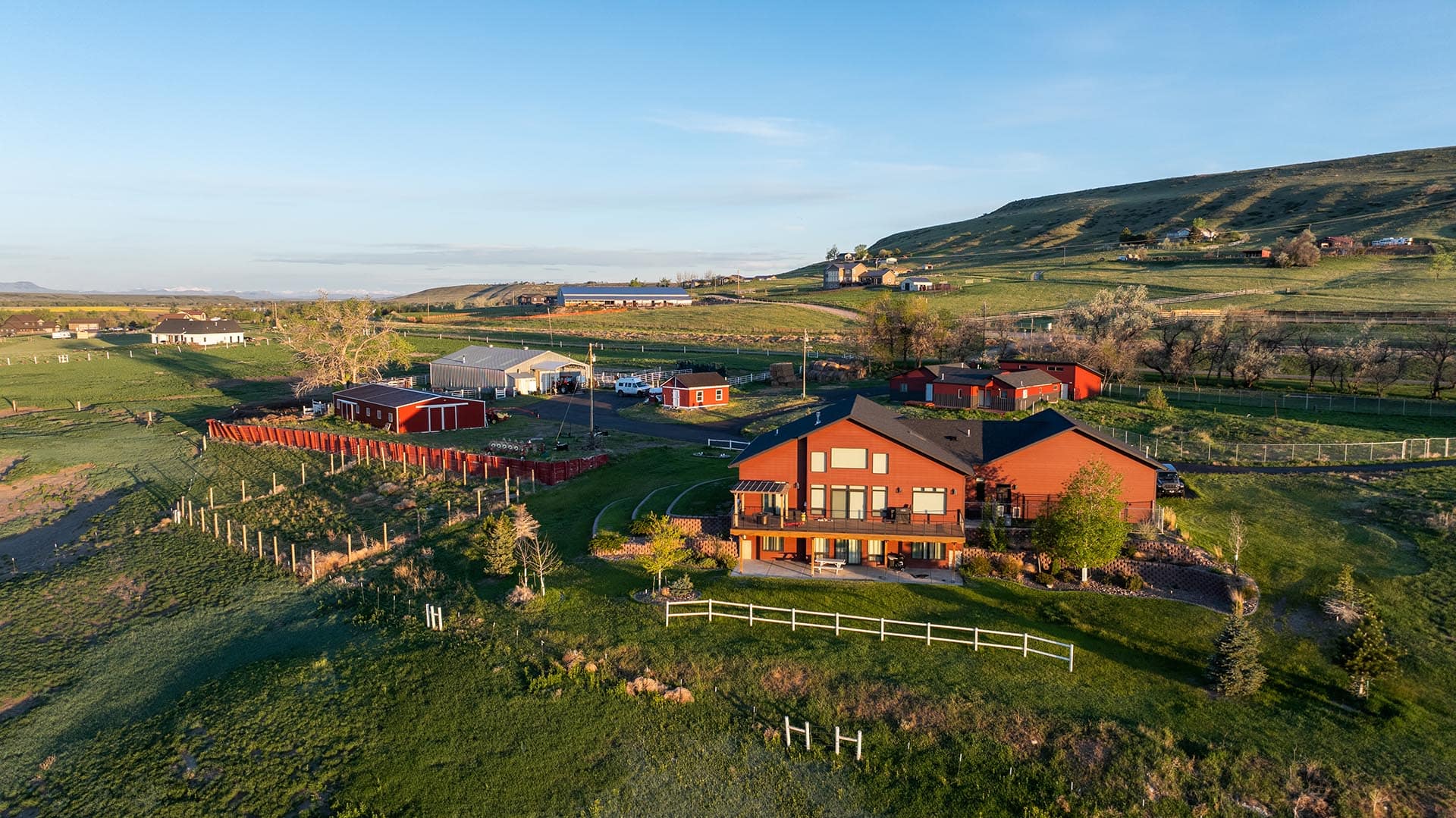 Aerial view of a rural farmstead with red houses, red barns, white fences, and green pastures under a clear blue sky.