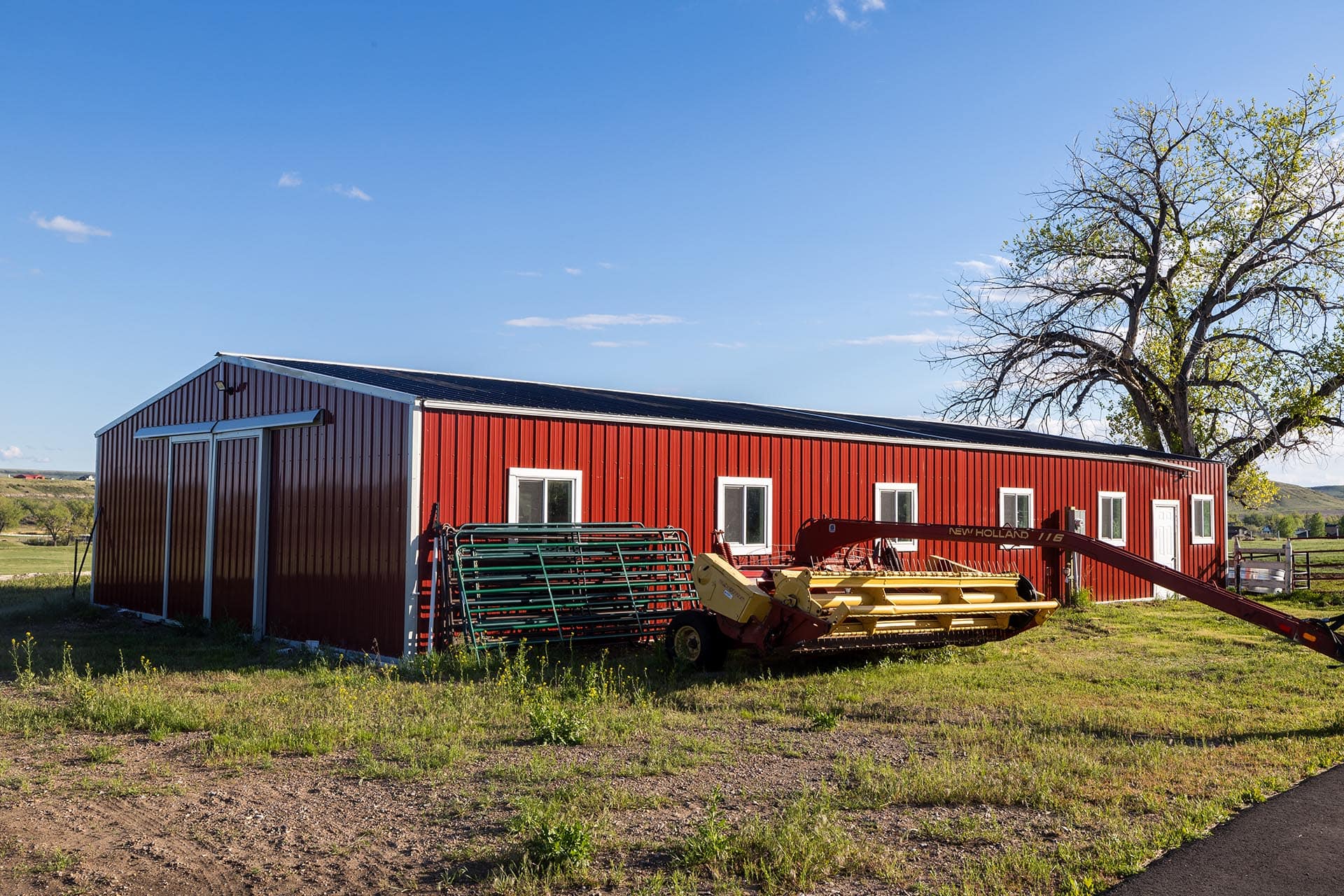 Red barn with white trim and farming equipment outside, under a clear blue sky with a large tree on the right.