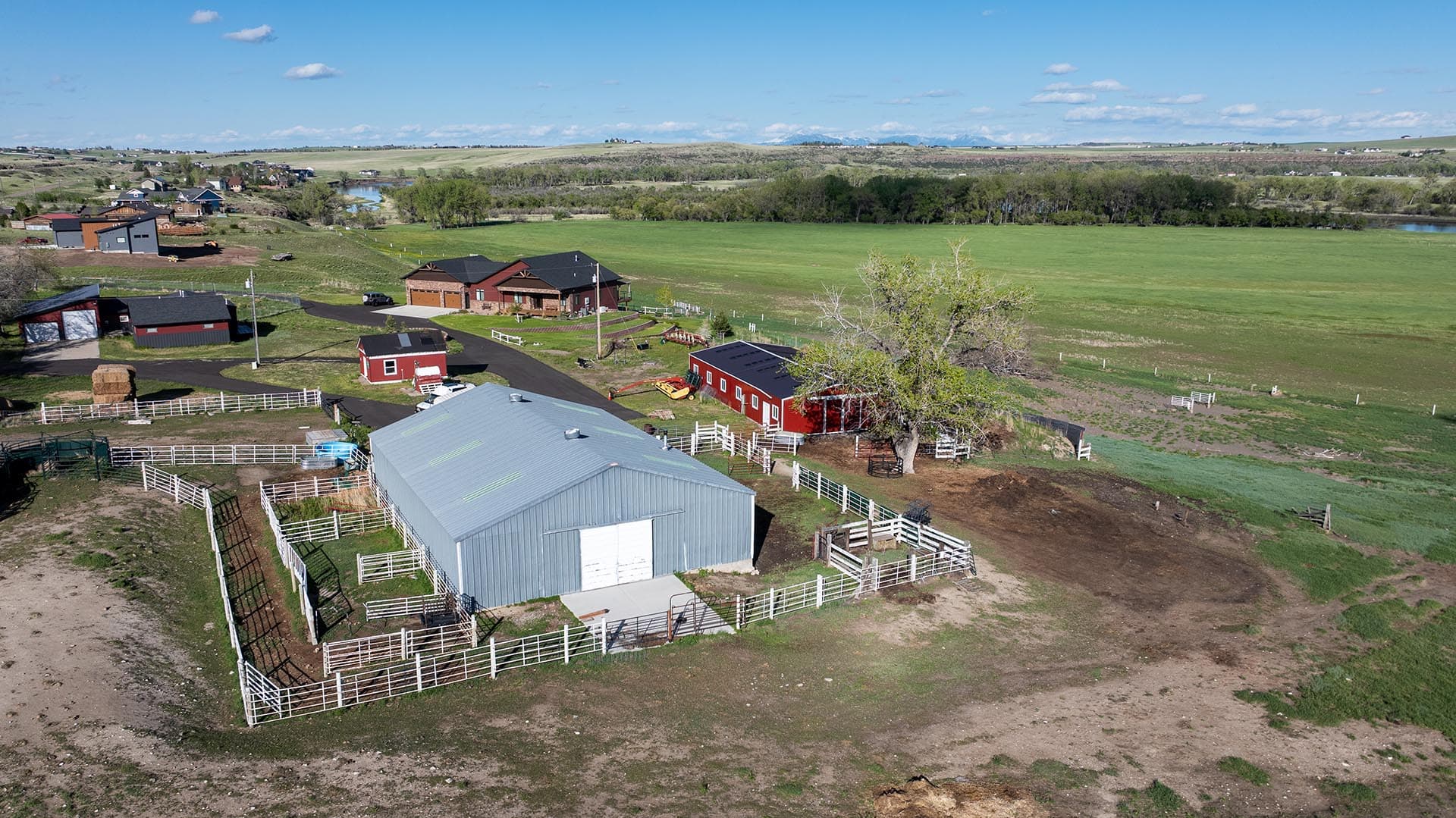Aerial view of a farm with red barn, smaller red shed, white fences, and a large gray metal barn amid green pastures and blue sky.