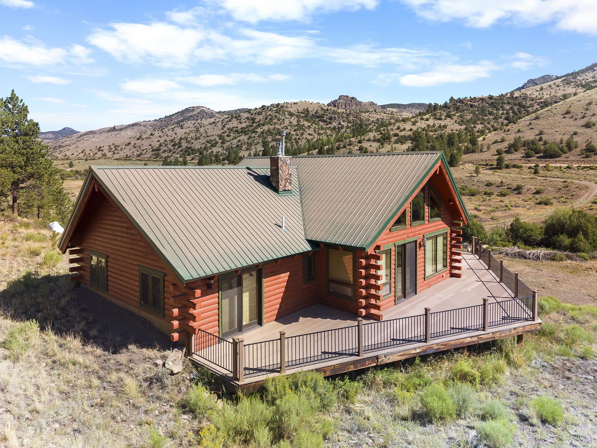 Red log cabin with a green metal roof on a wooden deck, set in a dry hillside landscape under a blue sky.