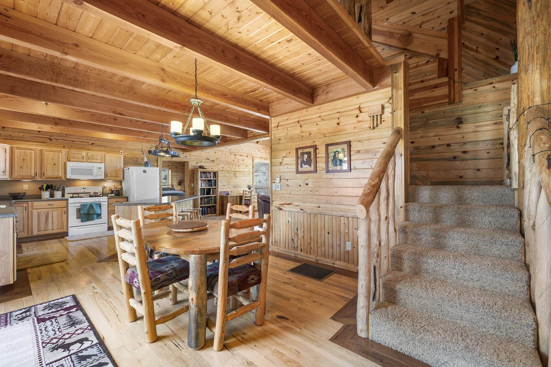 Rustic cabin interior with exposed wooden beams, a kitchen to the left, and a round wooden dining table in the center under a chandelier, plus carpeted stairs on the right