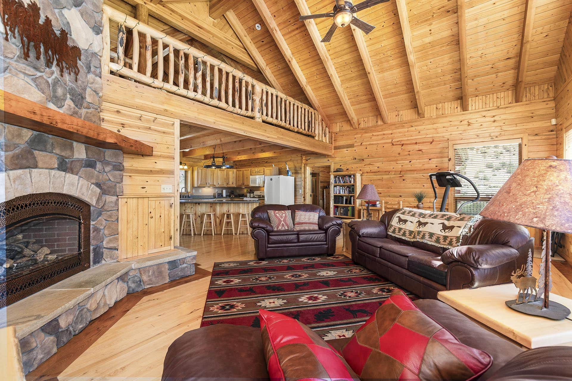 Rustic log cabin living room with a stone fireplace, wooden beams, loft railing, and leather sofas.