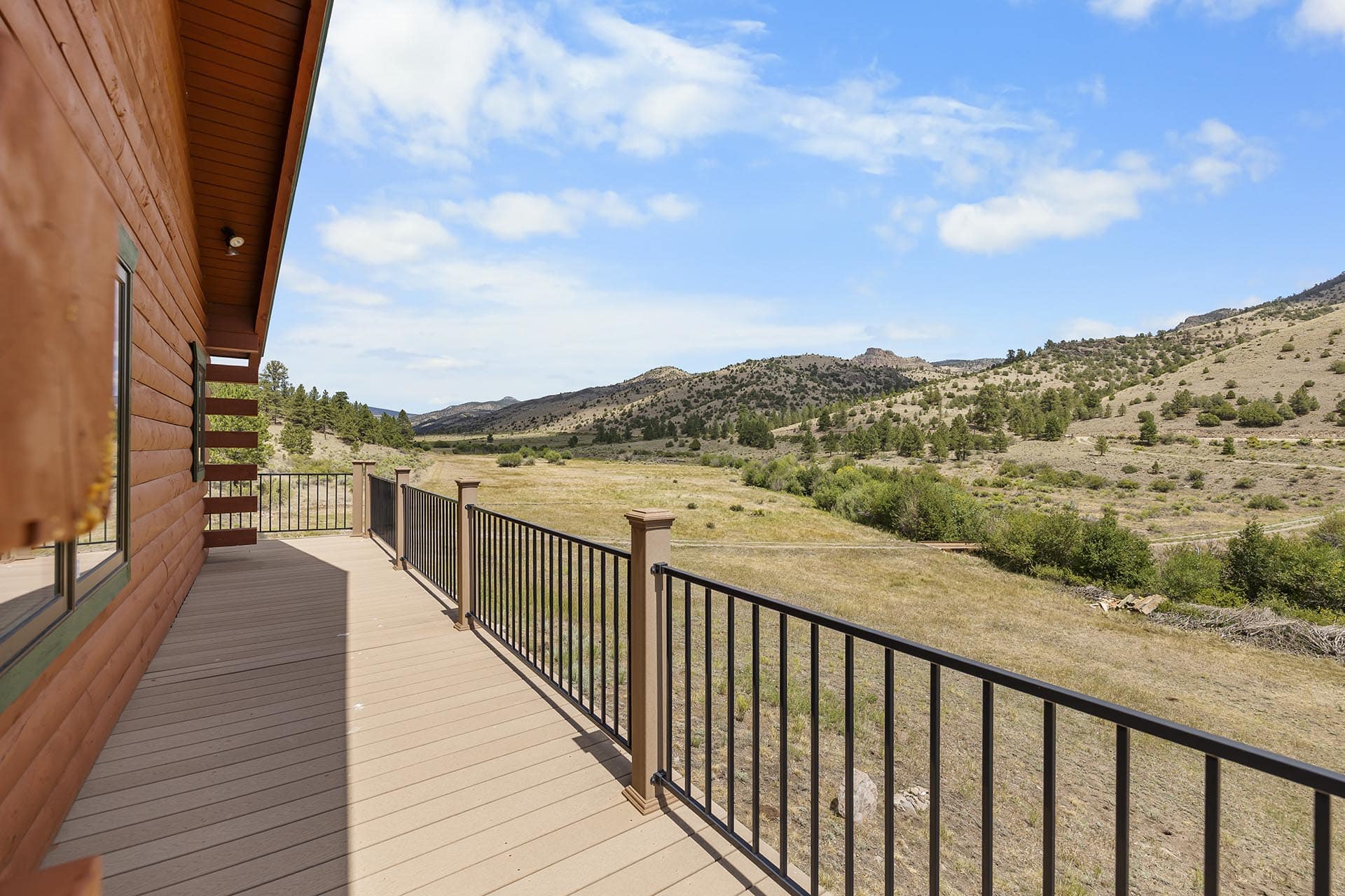 Wooden cabin deck with black railing overlooking a grassy valley and distant hills under a blue sky.