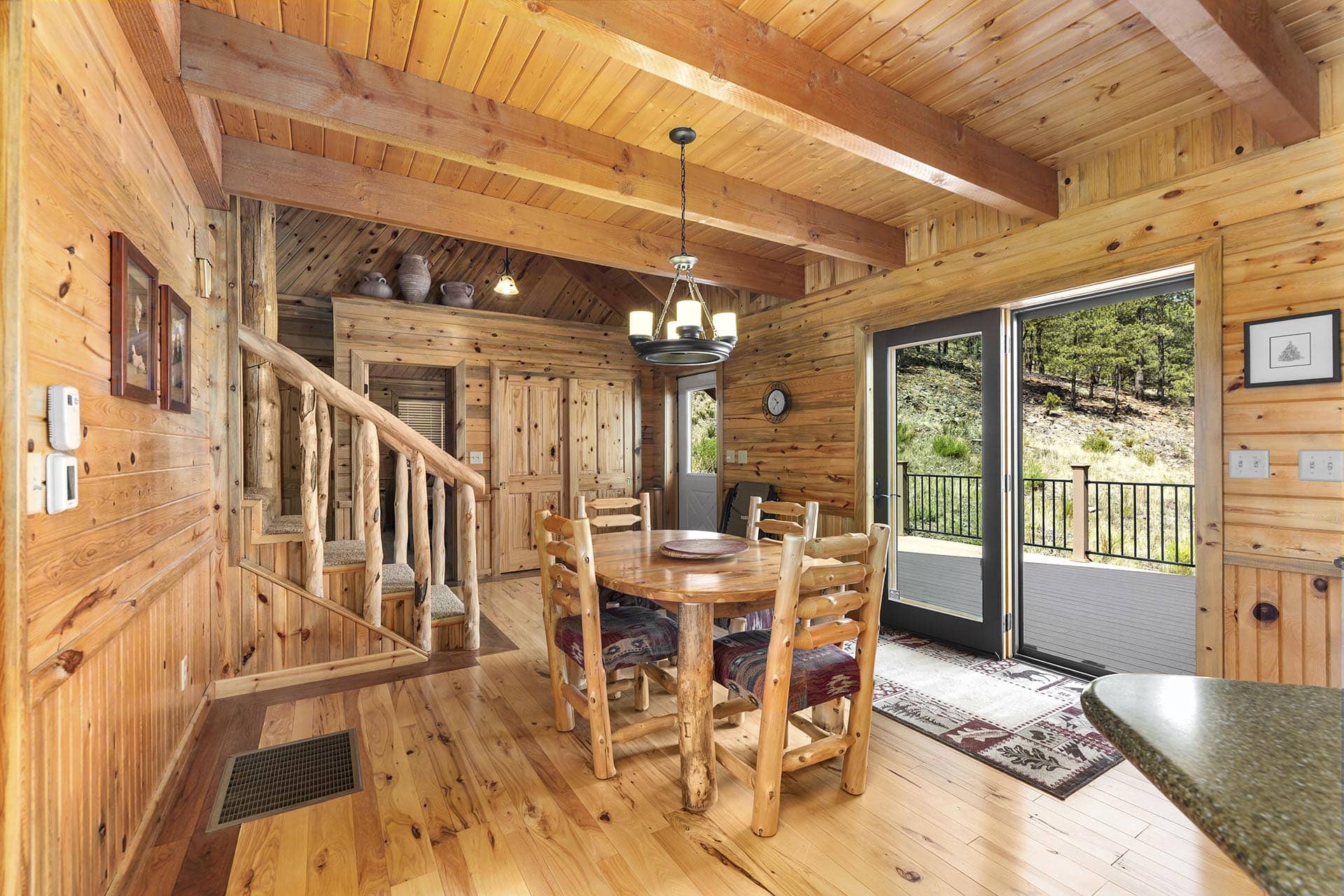 Rustic wooden dining area with a round table and log chairs inside a cabin, stairs to the left and large glass doors opening to a deck.