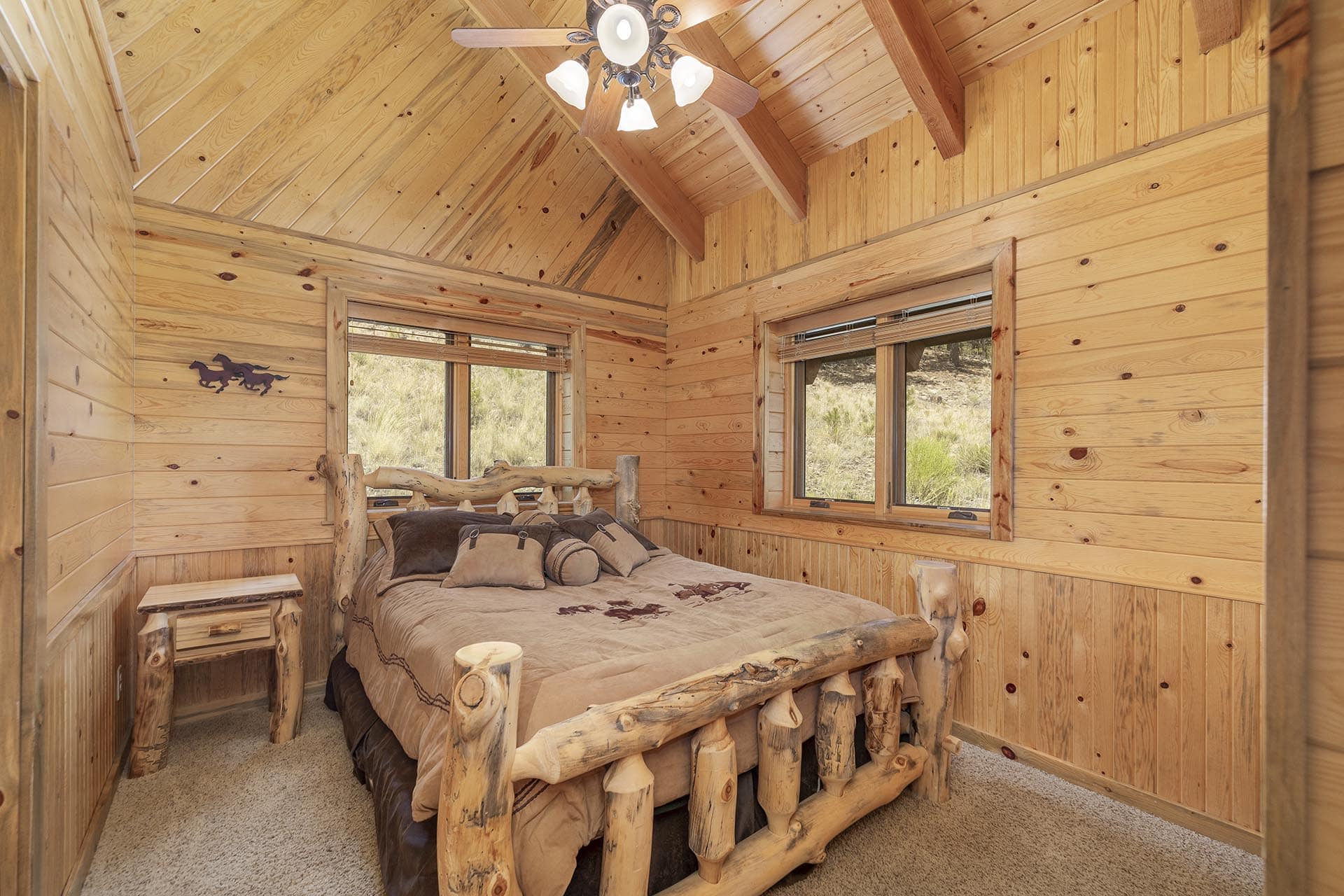 Rustic cabin bedroom with a log-style bed, wood-paneled walls, and two windows showing an outdoor view.