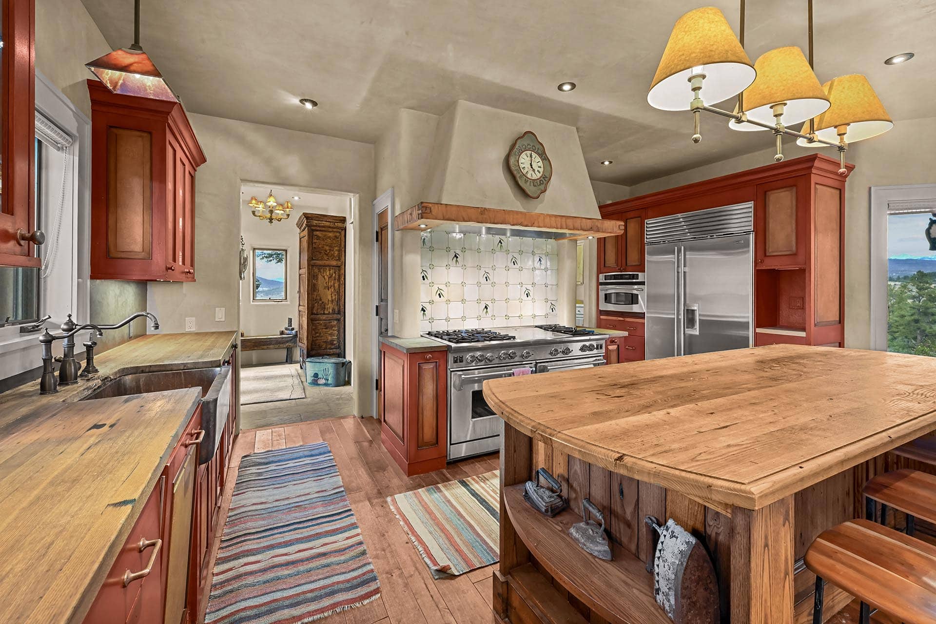Rustic kitchen with red cabinets, a large wooden island, and stainless steel appliances beneath warm pendant lighting.