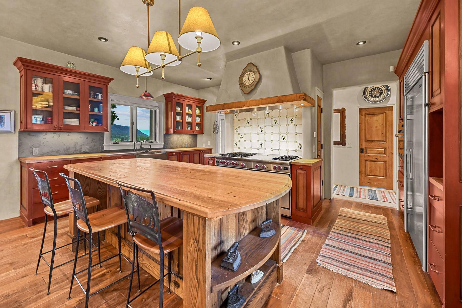 Kitchen with red wooden cabinets and a large wooden island, metal bar stools, and a stainless stove under a rustic wooden hood.