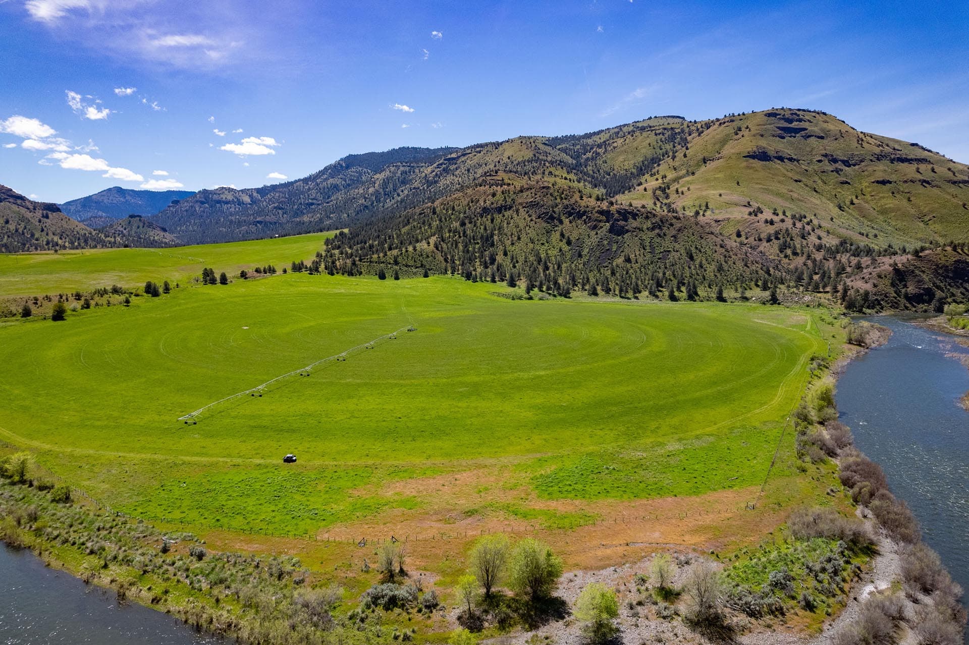 Aerial view of a green valley with rolling hills and a winding river along the right edge under a clear blue sky.