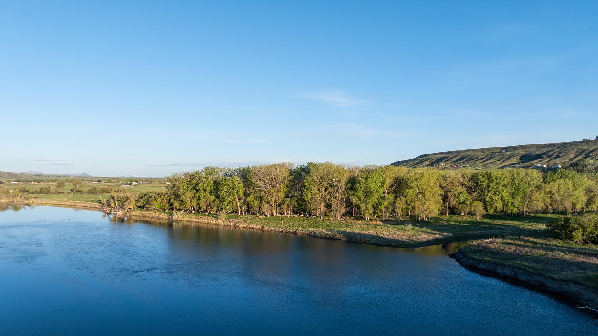Calm river with a tree-lined shore and grassy hill in the distance under a clear blue sky.