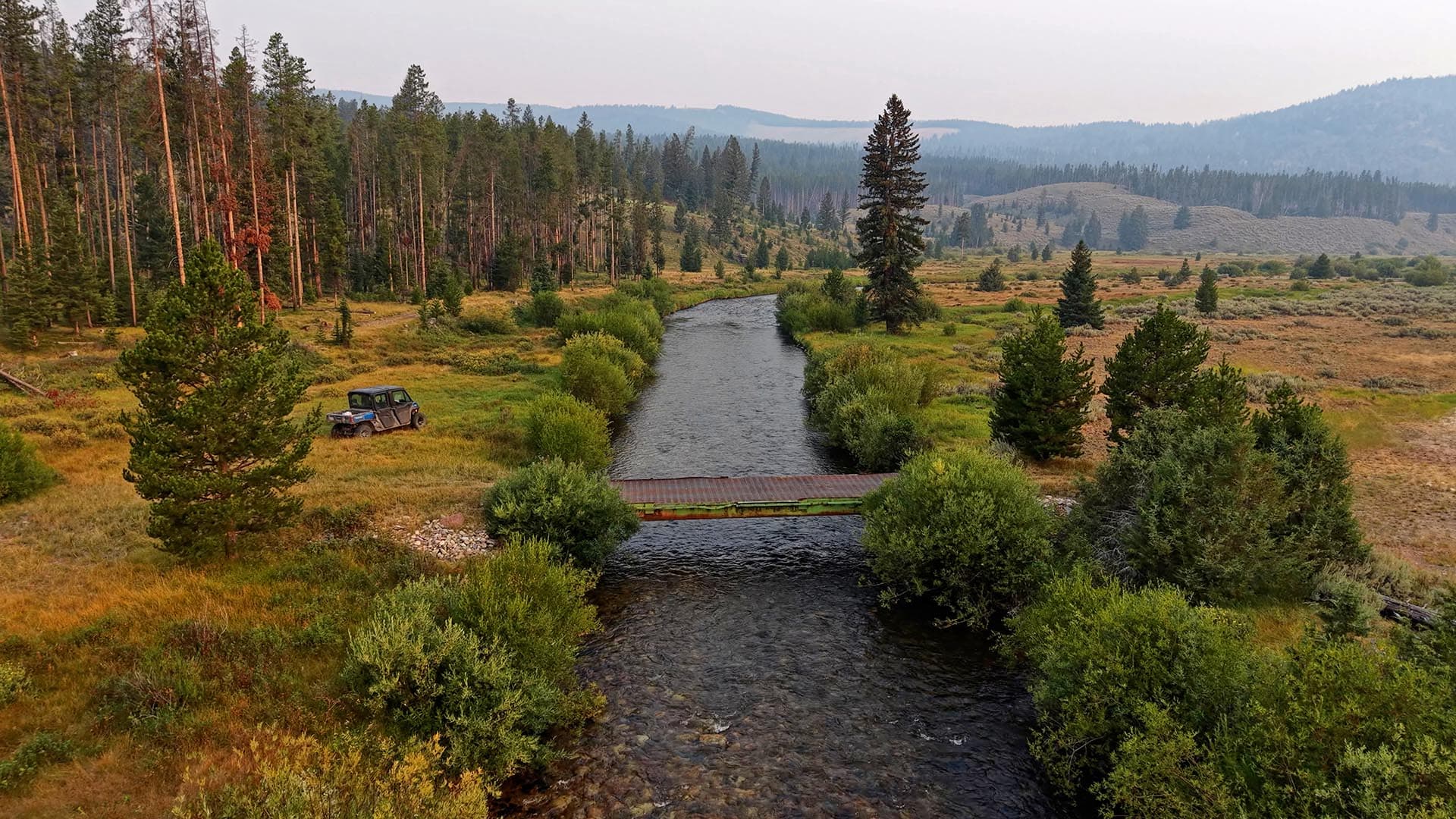 A shallow river runs through a grassy valley with a small wooden bridge; an off-road jeep sits on the left among pines and shrubs, with forested hills in the distance.