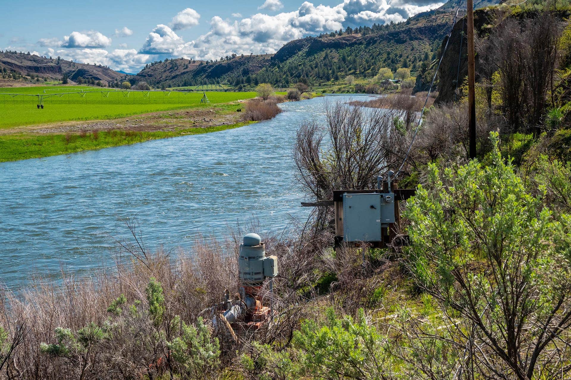 River runs through a grassy valley with distant hills and an irrigation sprinkler system across the field on the left side of the image.