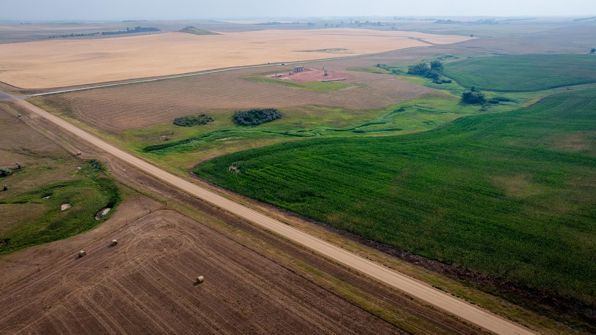 Aerial view of farmland with a diagonal dirt road, hay bales, and patchwork fields in brown and green.