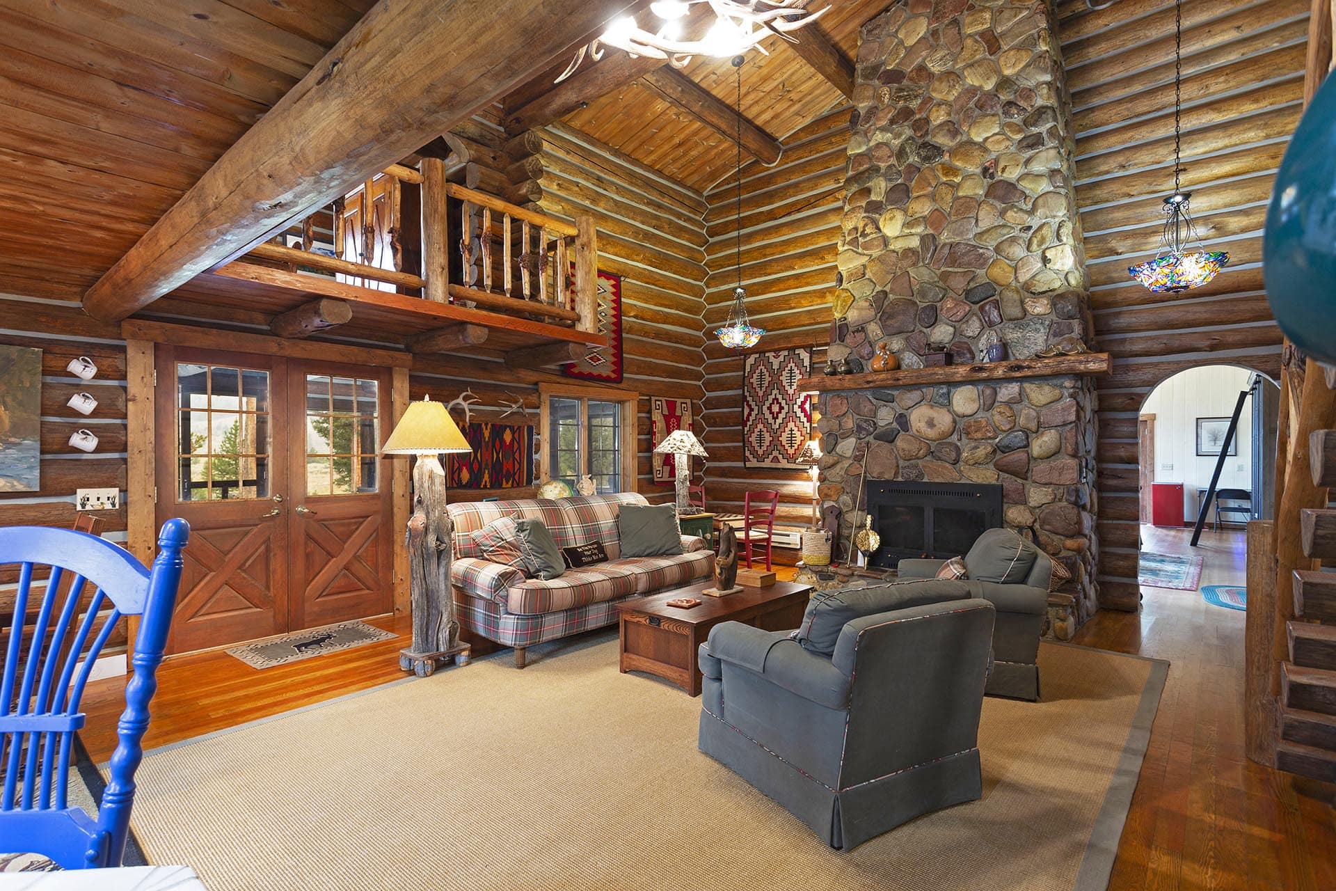 Cozy rustic log-cabin living room with a stone fireplace, exposed wood beams, and a loft balcony over plaid sofas and chairs.