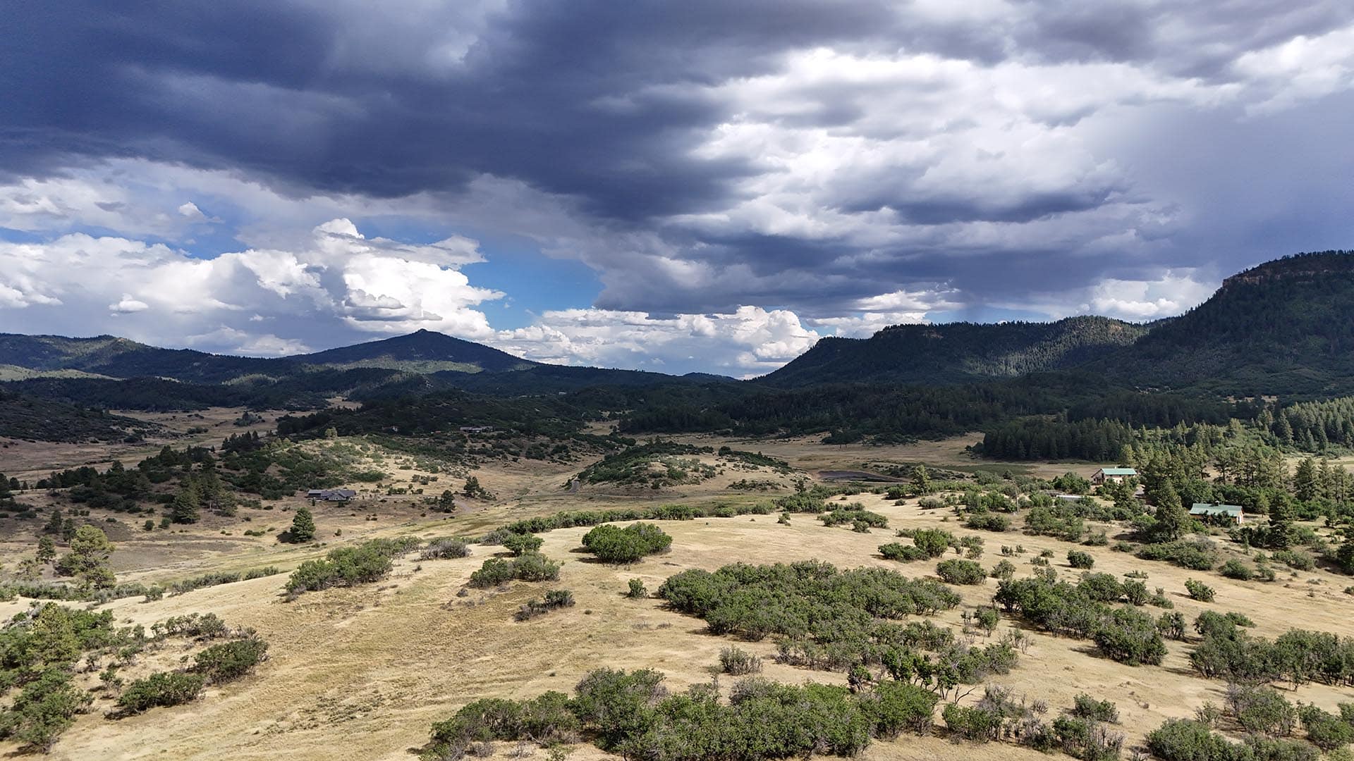 Wide valley with rolling hills and scattered shrubs under dark storm clouds looming over distant mountains; a few buildings with green roofs dot the landscape.