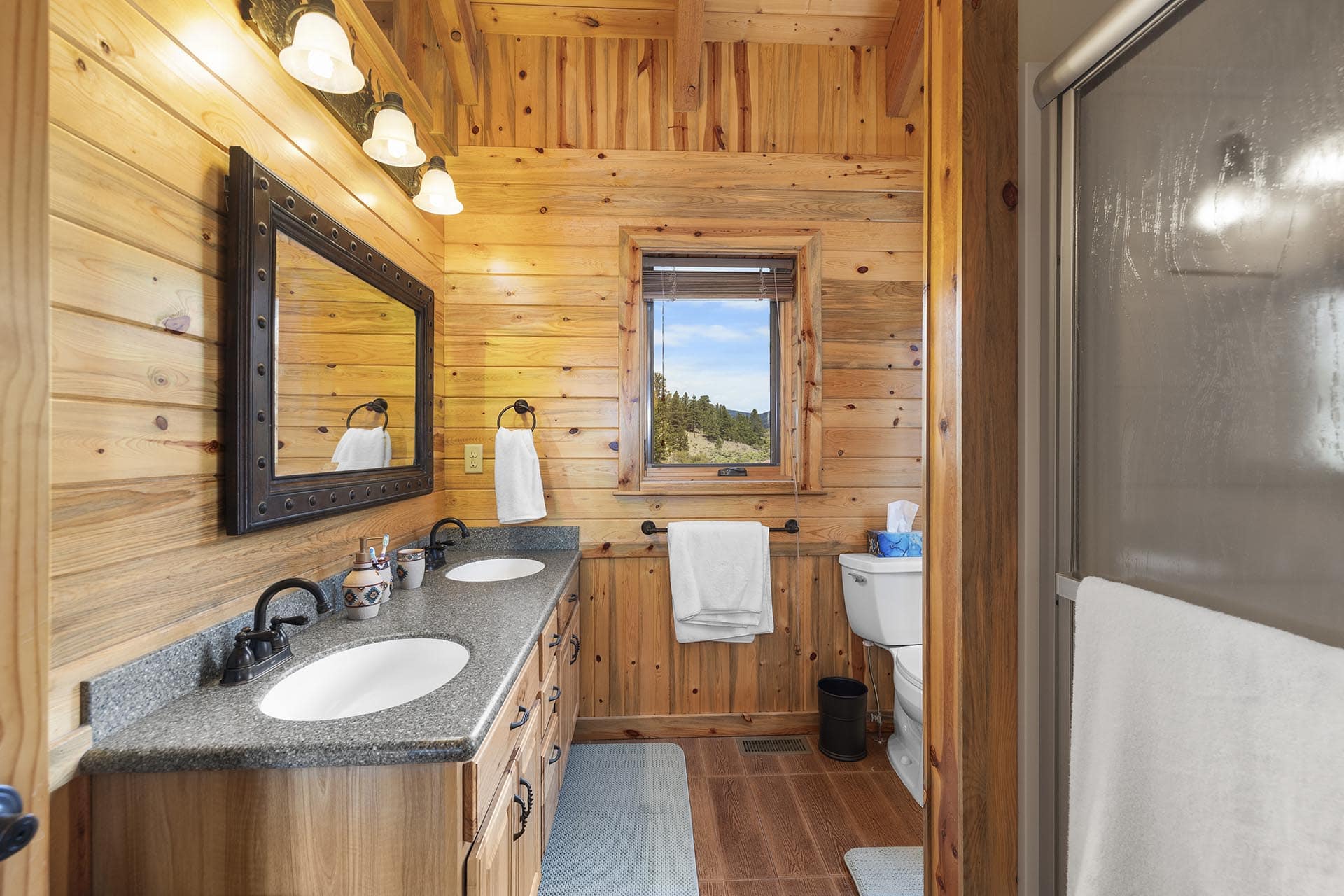 Rustic cabin bathroom with a double-sink vanity, black faucets, granite countertop, and a dark framed mirror on wood-paneled walls; window view of hills outside.