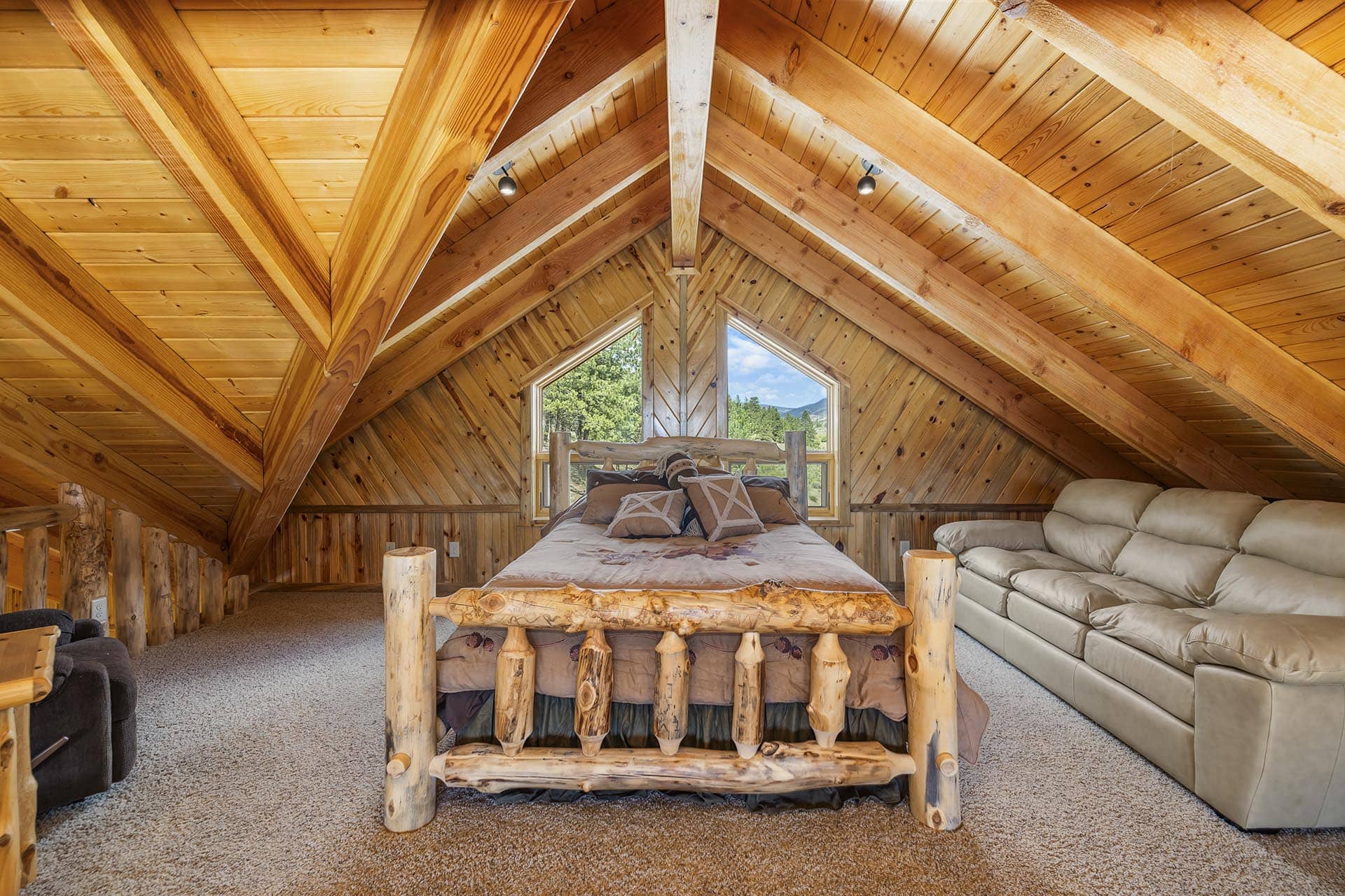 Rustic attic bedroom with a log bed, exposed timber beams, and a large window showing trees outside and mountains beyond.