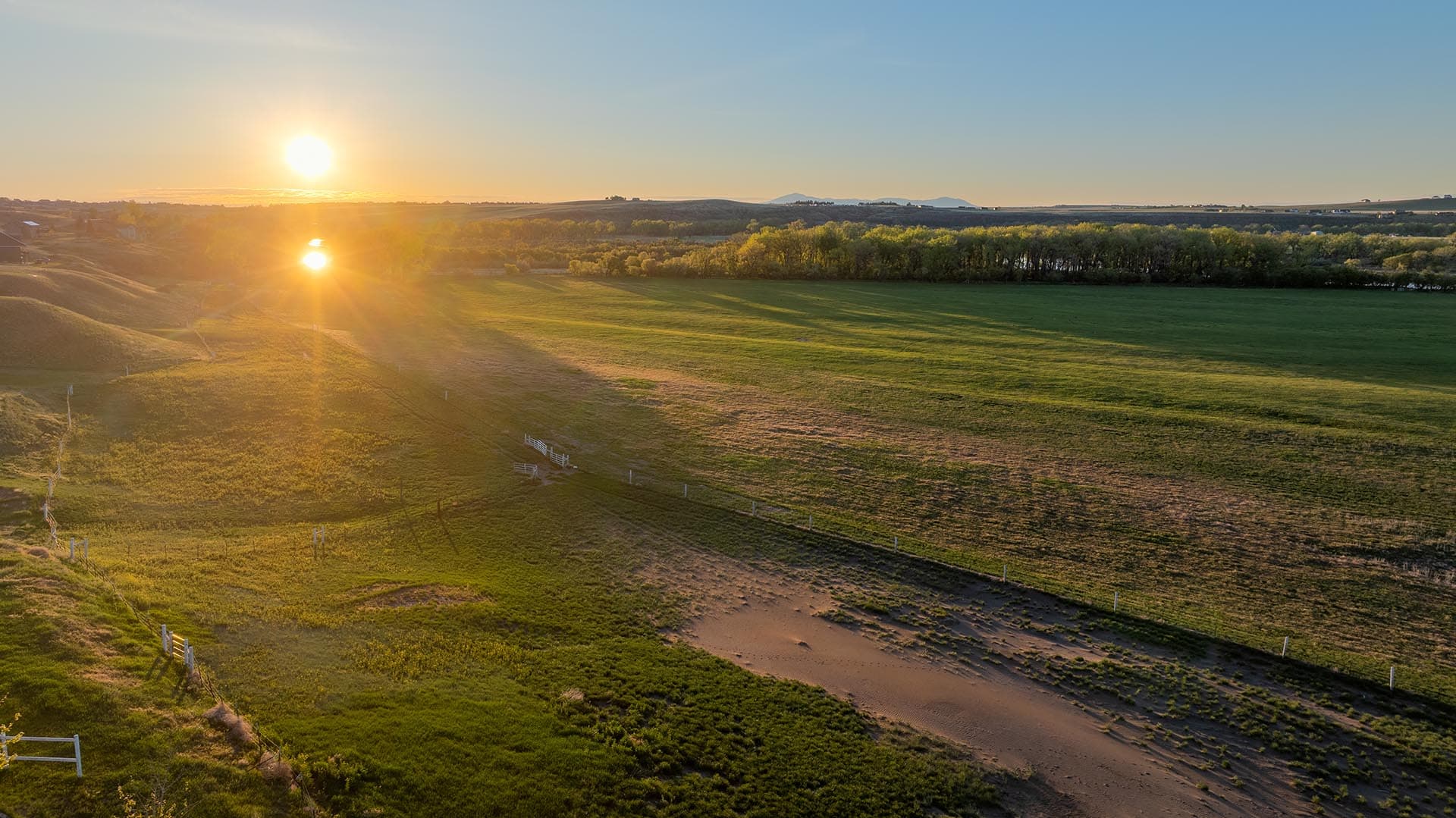 Aerial view of a rural landscape at sunset: fields, scattered fences, and a line of trees along the horizon with the sun low in the sky.