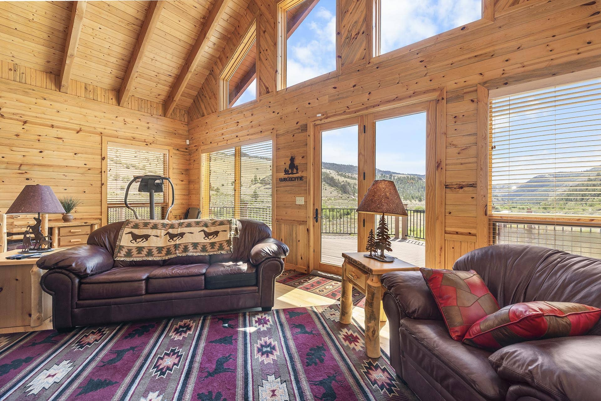 Cozy cabin living room with leather sofas, wooden walls, and a colorful geometric rug; large windows overlook mountains outside