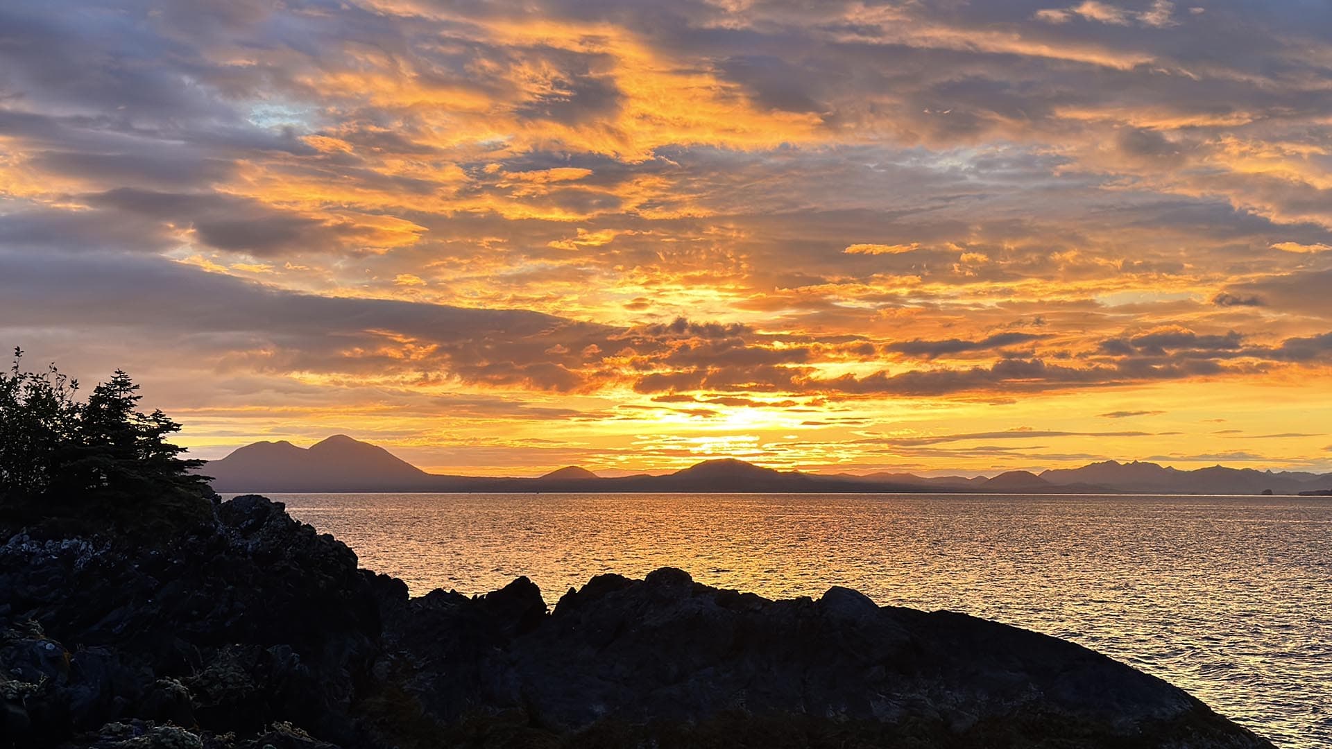 Sunset over calm water with orange clouds, silhouetted rocky shoreline in the foreground and distant island silhouettes.