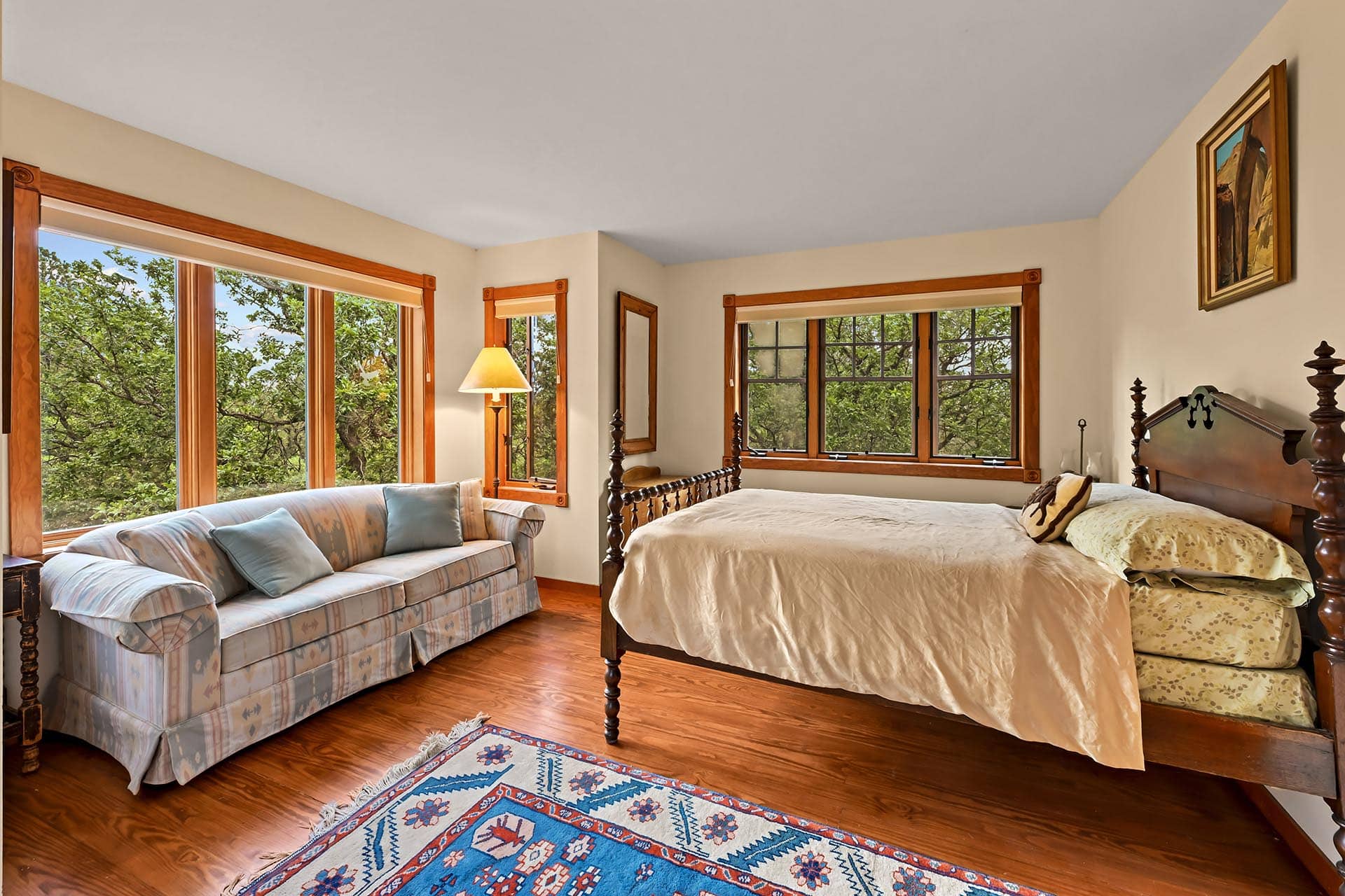 Cozy bedroom with a wooden four-poster bed, beige bedding, and a blue patterned rug by large windows looking onto green trees