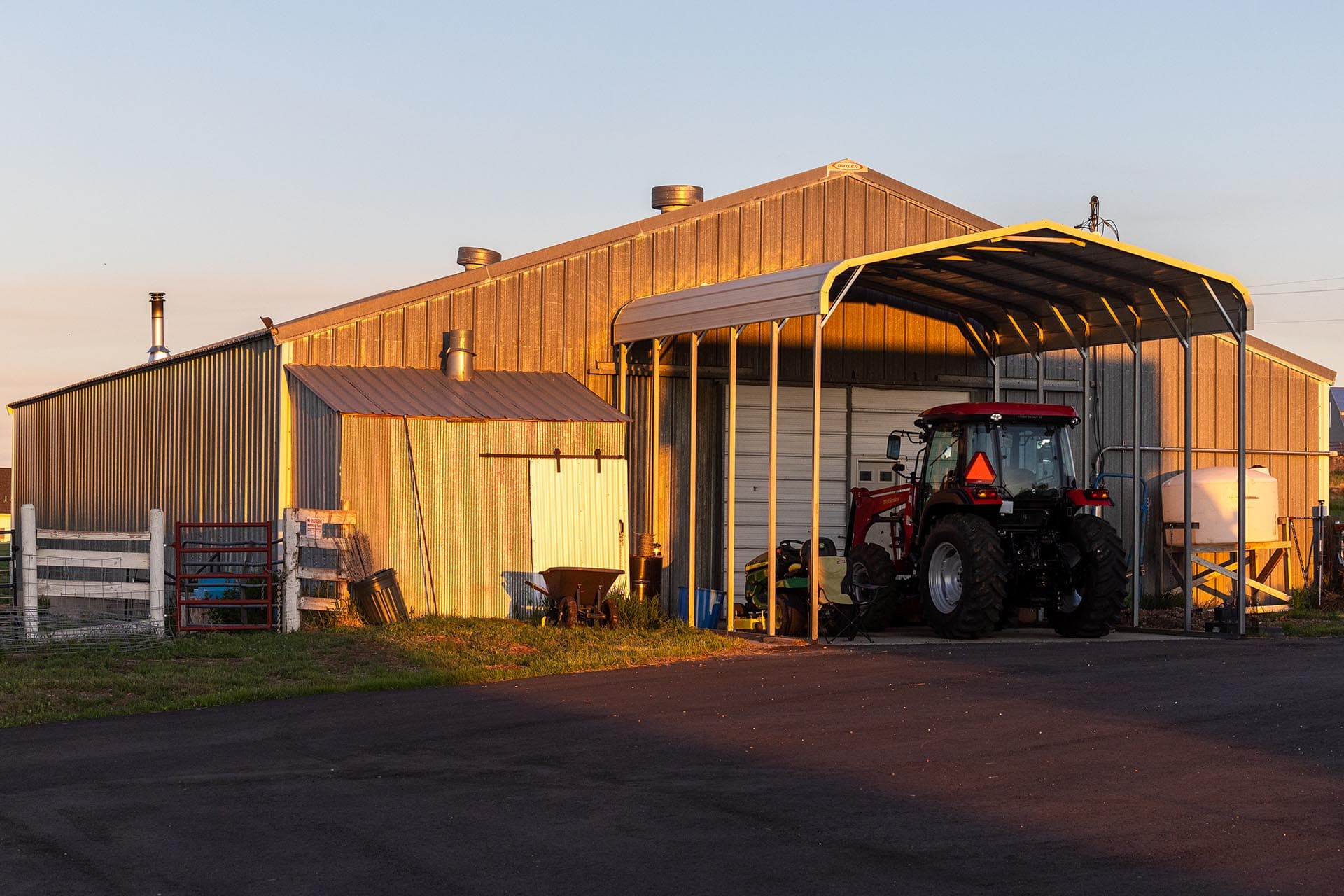 Golden-hour view of a metal barn with a covered area housing a red tractor parked inside