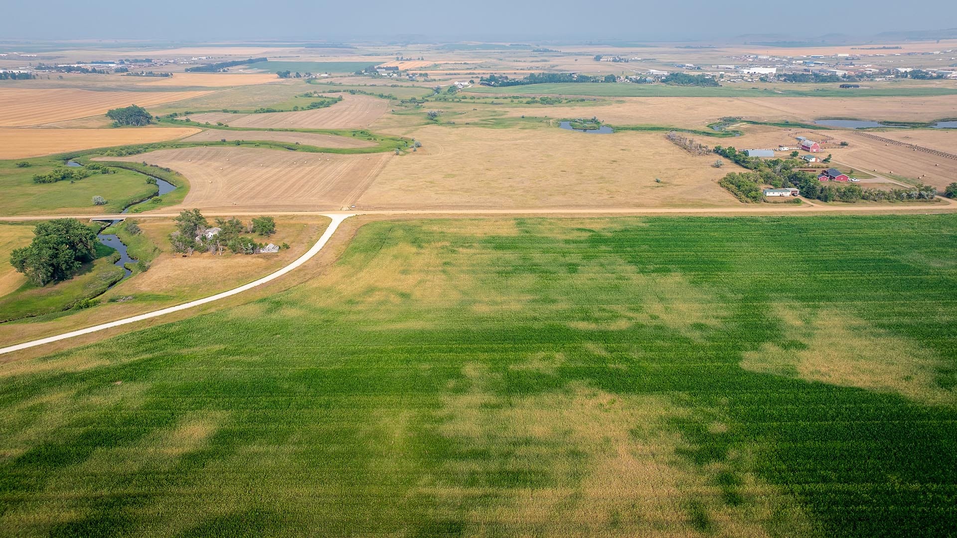 Aerial view of farmland with a winding dirt road, green fields and distant farm buildings in the countryside.