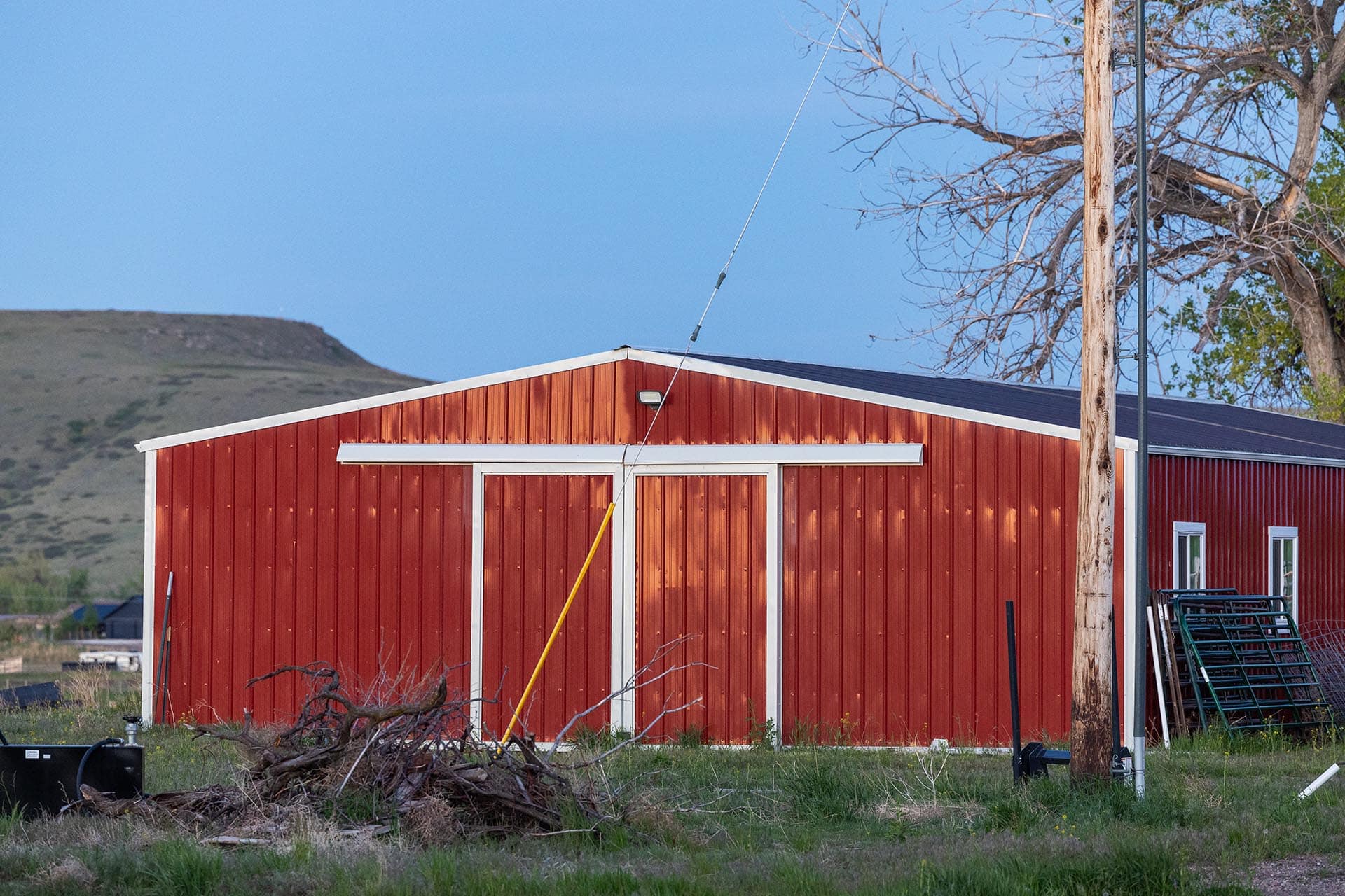 Red metal barn with white trim in a rural yard, a yellow pole leaning nearby, and a bare-branched tree to the right against a blue sky.