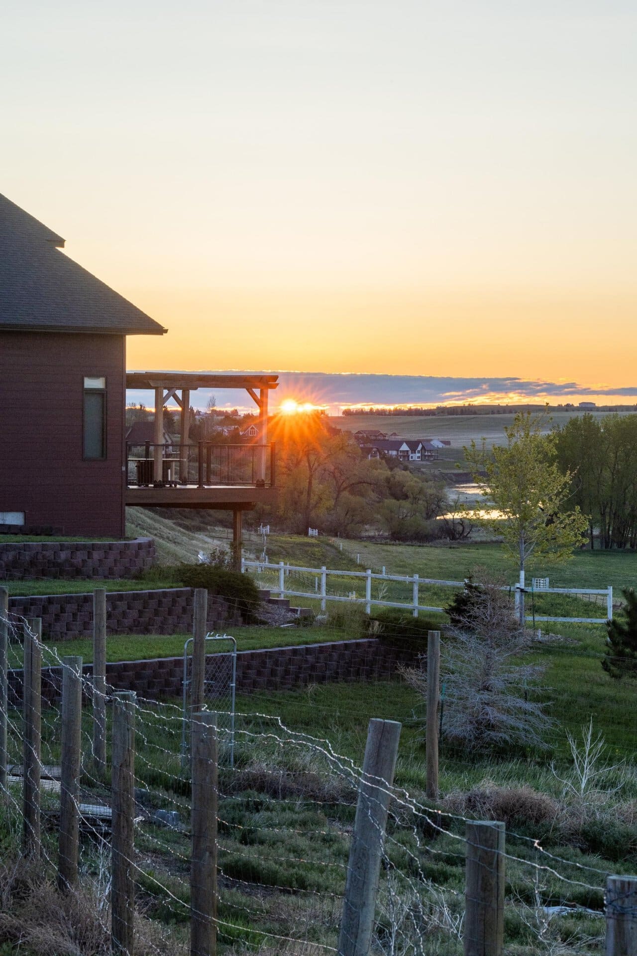 Sunset over a rural yard with a wooden house and deck on the left, barbed-wire fence in the foreground, and fields extending to the horizon.