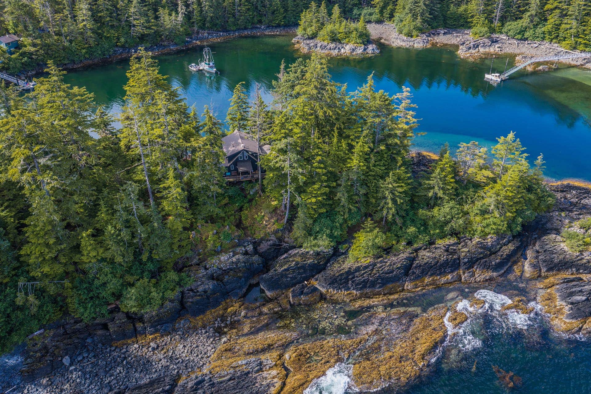 A cabin tucked among tall evergreen trees beside a turquoise lake with boats anchored nearby and rocky shoreline.