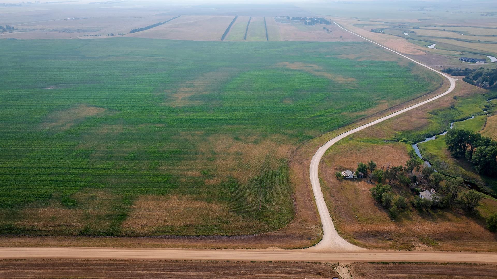 Aerial view of green farmland with a winding dirt road curving from bottom right, small farm buildings at the edge, and a narrow canal along the right side.