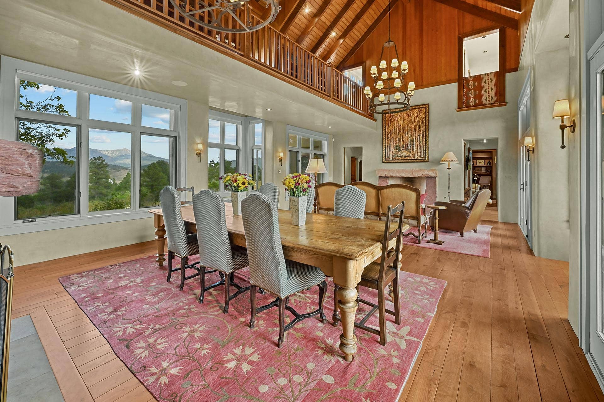 Bright dining room with a rustic wooden table and upholstered chairs, pink patterned rug, and large windows showing a mountain landscape; a wooden mezzanine runs along the upper level.