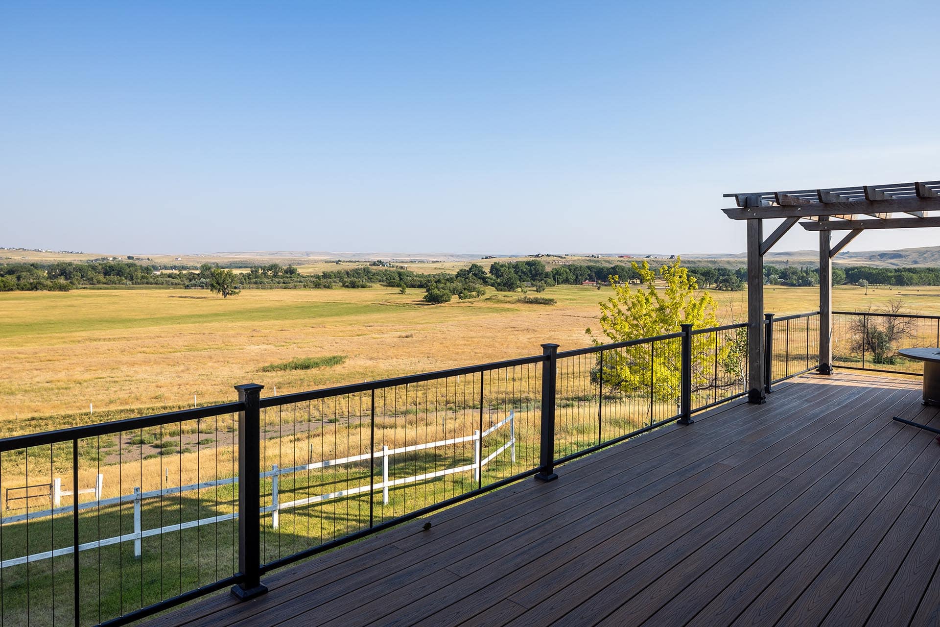 View from a wooden deck with metal railing overlooking sunlit golden fields and distant trees under a clear blue sky.