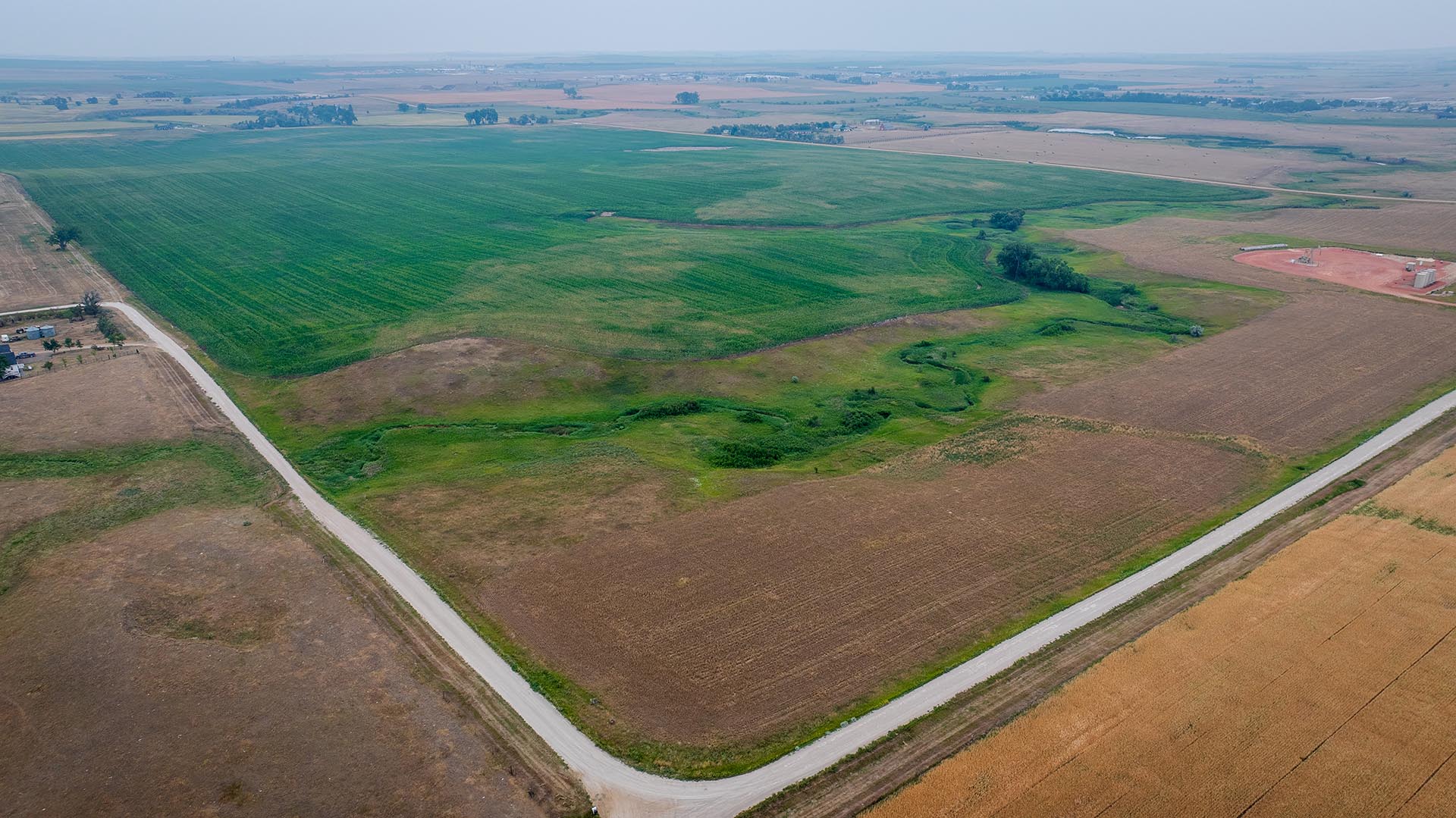 Aerial view of farmland with green crop fields and brown tilled plots, intersecting roads forming a corner in the foreground.